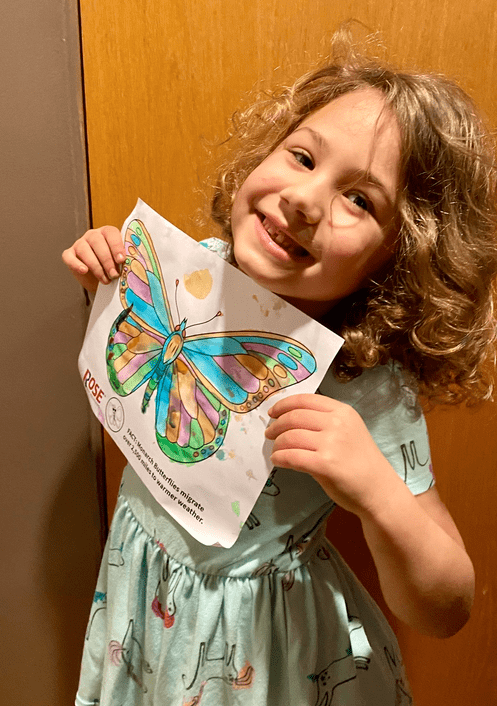 A young child with curly hair smiles and holds up a colorful, hand-colored butterfly drawing while standing indoors, proudly dressed in a northwestern football shirt.