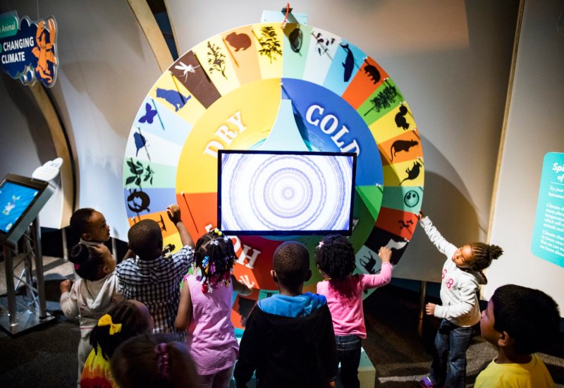 042816 NatureMuseum 1204 A group of children gather around a colorful spinning wheel display in a nature museum exhibit sponsored by Rose.