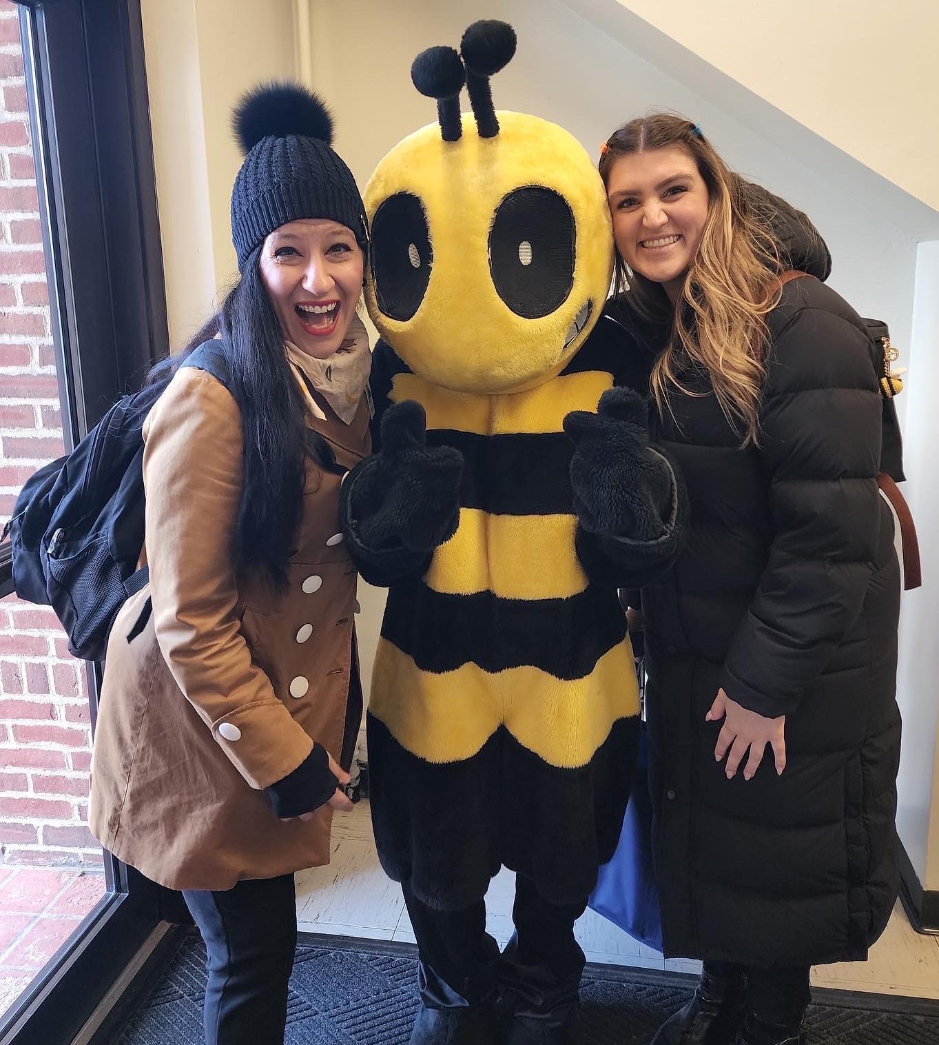 DC172C07 FF72 4627 84D4 51F4416E8181 Two women stand on either side of a person in a bee costume, all smiling indoors near a window and a brick wall—capturing the fun spirit often found at About Rose Pest Solutions events.