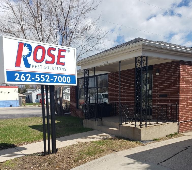 Brick building with a black railing and sign reading "Rose Pest Solutions" and a phone number in front; trees and other buildings in the background, showcasing Racine pest control services.